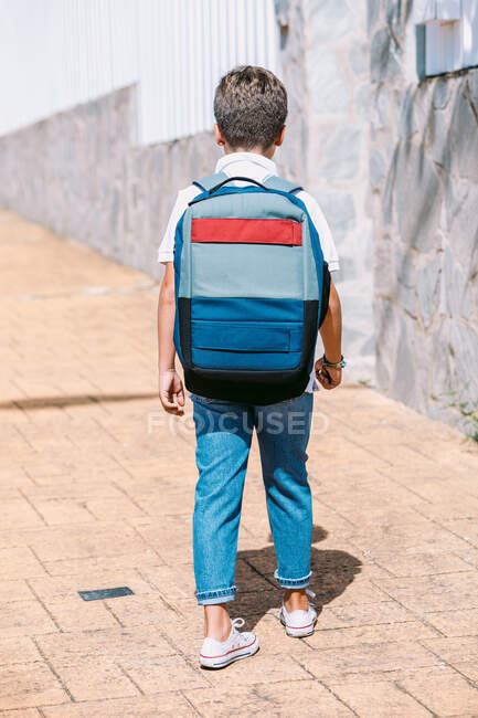 Back view of unrecognizable schoolchild in ripped jeans and gumshoes on tiled pavement in sunny town — Stock Photo