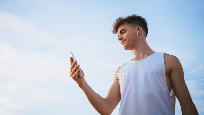 From below of young male athlete in undershirt with cellphone listening to music from true wireless earbud under cloudy sky — Stock Photo