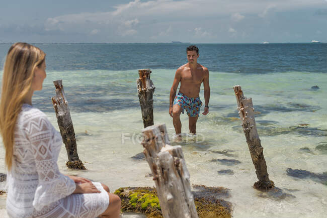 Красивый парень с загорелым мускулистым туловищем разговаривает с очаровательной молодой женщиной в пляжной одежде во время романтического медового месяца на тропическом побережье — стоковое фото