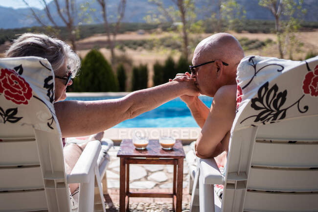 Вид ззаду без сорочки люблячий старий чоловік цілує руку дружини, насолоджуючись літнім днем на шезлонгах на узбережжі басейну — стокове фото