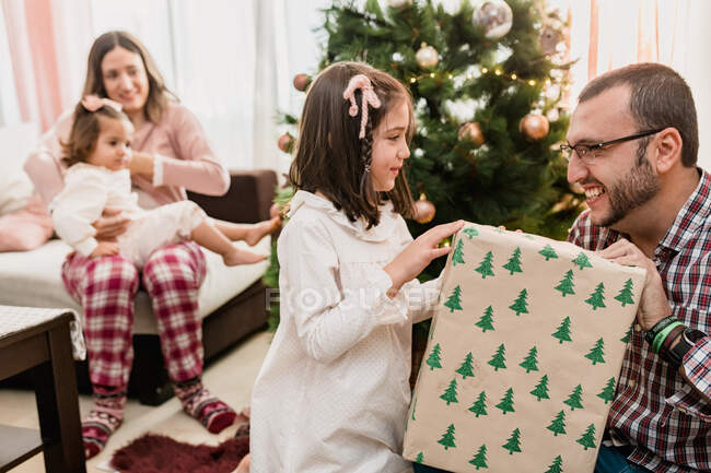 Pai alegre passando caixa de presente para menina contra esposa com criança durante o feriado de Ano Novo em casa — Fotografia de Stock