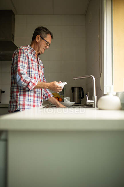 Vista laterale di calma felice maturo maschio lavaggio piatti sporchi mentre in piedi vicino lavello in cucina e fare i lavori domestici — Foto stock