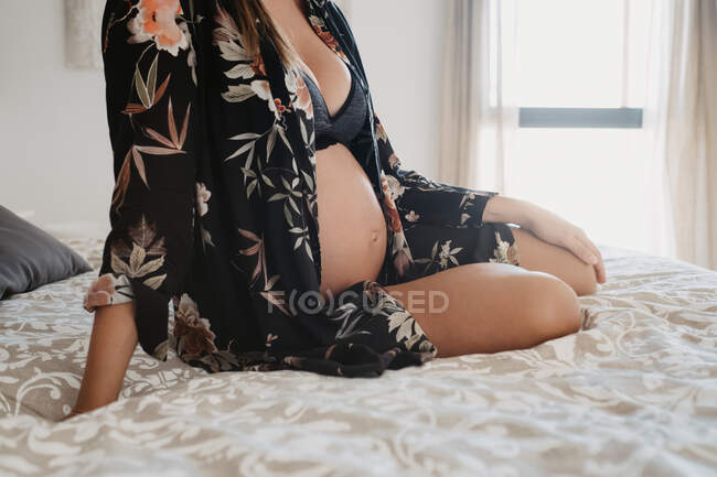 Cultivado mujer embarazada irreconocible en ropa interior sentado mirando hacia abajo en la cubierta de cama suave en casa - foto de stock