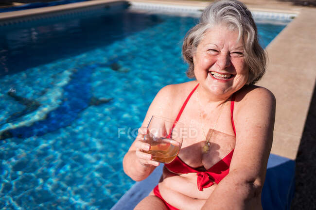 Высокий угол восторженной пожилой туристки в бикини, ярко смеющейся, расслабляясь у бассейна с напитком — стоковое фото