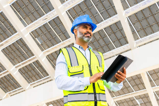 Bajo ángulo de ingeniero masculino hispano de mediana edad en uniforme con la tableta mirando hacia otro lado bajo la estación de energía solar - foto de stock