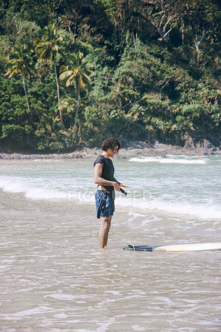 Side view of young sportsman with leash and surfboard in wavy ocean against exotic trees in sunlight — Stock Photo