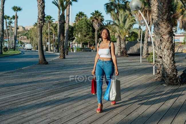 Jovem compradora afro-americana alegre com sacos de compras olhando embora enquanto caminhava na rua — Fotografia de Stock
