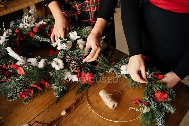 Copped unrecognizable female friends standing at wooden table making creative Christmas wreath from decorative fir and red ribbons for holiday celebration — Stock Photo