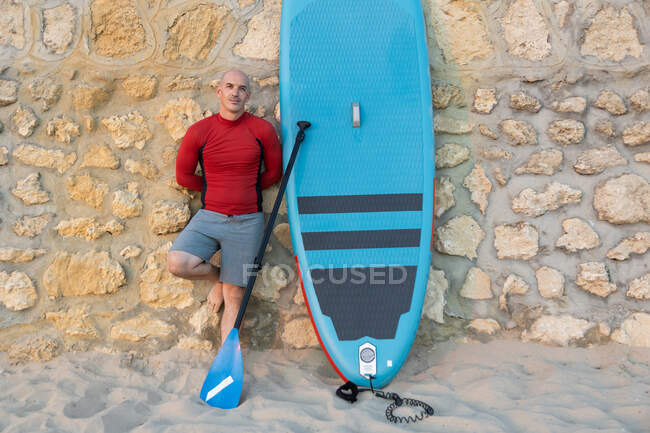 Hombre surfista en traje de neopreno y sombrero de pie mirando a la cámara con paleta y tabla SUP mientras se prepara para surfear en la orilla del mar apoyado en la pared de piedra - foto de stock