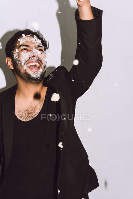 Hombre feliz con la cara sucia riendo con la boca abierta mientras vomita pastel de cumpleaños roto en el estudio - foto de stock