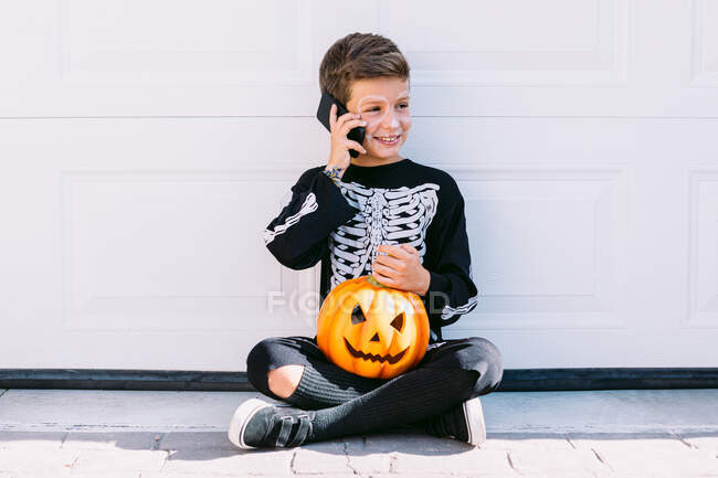 Повне тіло веселого хлопчика в чорному костюмі скелета з розфарбованим обличчям і різьбленим гарбузом на Хеллоуїн розмовляє на мобільному телефоні, сидячи біля білої стіни на вулиці — стокове фото