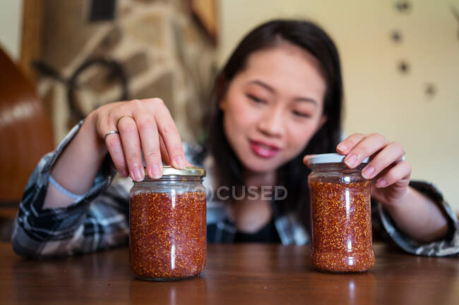 Jovem fêmea étnica com frascos de vidro de deliciosa marmelada de figo na mesa em casa no fundo borrado — Fotografia de Stock