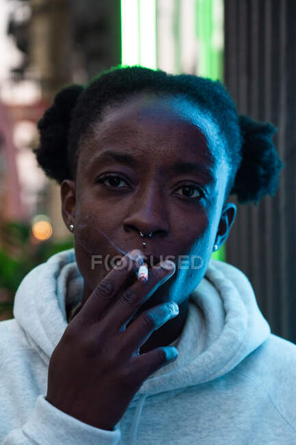 Pensativo afroamericana no binaria mujer en casual sudadera con capucha fumar cigarrillo y mirando hacia otro lado pensativamente - foto de stock