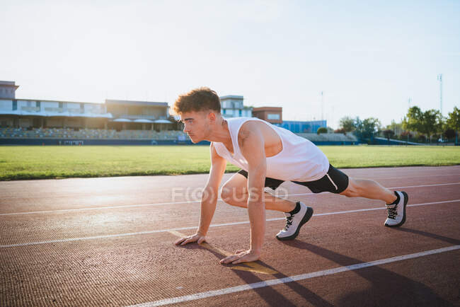 Вид сбоку спортсмен в кроссовках, стоящий в стартовой позиции перед тренировкой на трассе при солнечном свете — стоковое фото