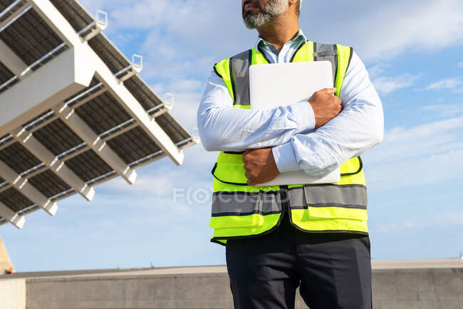 Низький кут обрізаного невпізнаваного зрілого етнічного чоловіка-інспектора в жилеті з буфером, який дивиться далеко, стоячи біля сонячної електростанції — стокове фото