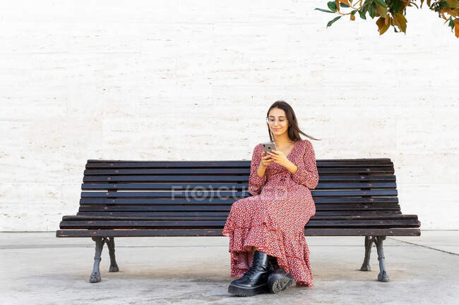 Full body of positive female in stylish clothes browsing smartphone and sitting on wooden bench on street in daytime — Stock Photo
