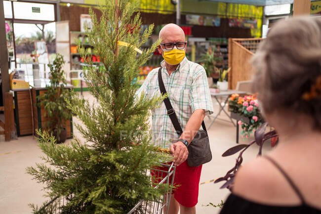 Seniorenkaufmann in steriler Maske mit Nadelbaum im Einkaufswagen sieht Frau in Gartengeschäft unkenntlich — Stockfoto