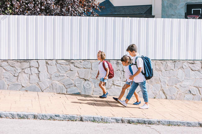 Vista lateral del colegial con mochila hablando con amigas mientras pasea sobre pavimento de baldosas contra pared de piedra a la luz del sol - foto de stock
