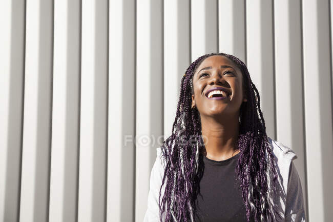 Happy young African American female with long dyed braids smiling brightly and looking up while having fun against gray wall in sunlight — Stock Photo