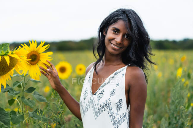 Mulher étnica adulta sincera olhando para a câmera no prado tocando flores florescentes no campo no fundo borrado — Fotografia de Stock