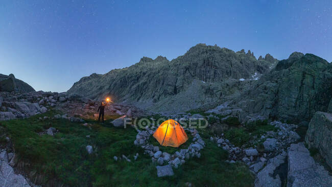 Picturesque scenery of unrecognizable traveler standing with bright light in hand near tent placed among rocky mountains under cloudless night sky — Stock Photo