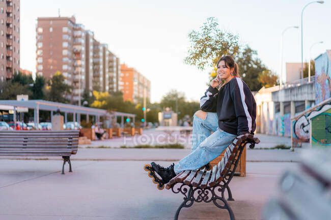 Cuerpo completo de una joven mujer positiva que usa sudadera con capucha negra casual y jeans azules claros con hendiduras y patines sentados en la parte posterior del banco de madera en el parque de skate - foto de stock