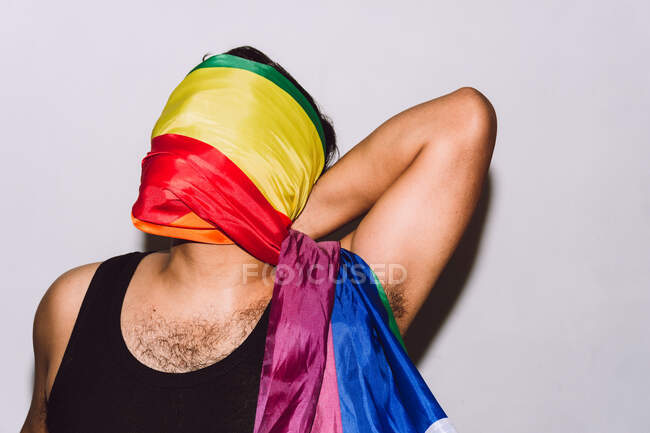 Hombre homosexual irreconocible con la cara envuelta en el símbolo de la bandera del arco iris de la comunidad LGBT contra el fondo blanco - foto de stock