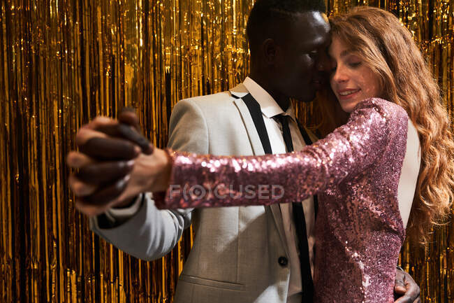 Jeune couple multiracial embrassant et tenant la main tout en dansant contre tinsel lors de la célébration du Nouvel An — Photo de stock