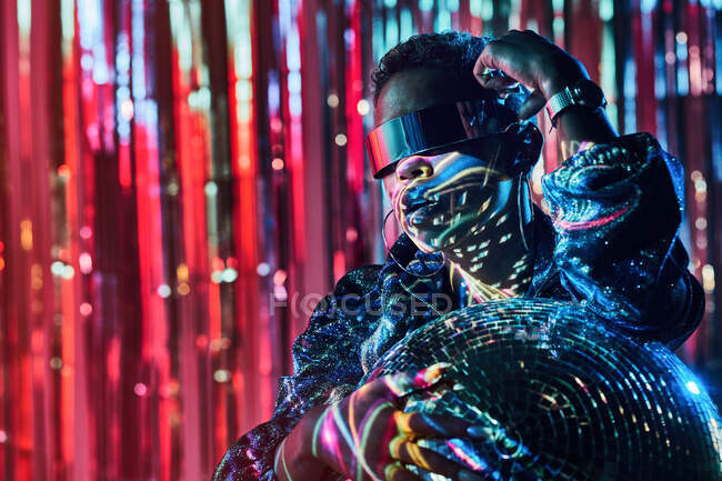 Mulher étnica na moda em óculos cyberpunk e relógio de pulso tocando bola disco em raio de luz — Fotografia de Stock