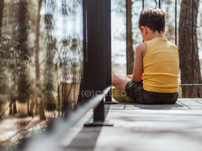 Вид сзади одинокого ребенка, сидящего на деревянной террасе дома в лесу в солнечный день — стоковое фото