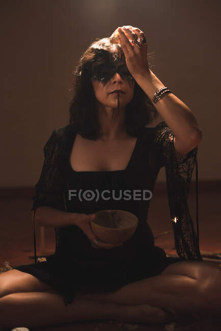 Dreamy enchantress with painted face and skull casting spell during mystic ritual in room with dim light with eyes closed — Stock Photo