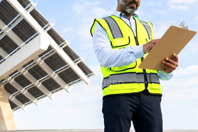 Low angle of cropped unrecognizable mature ethnic male inspector in vest with clipboard looking away while standing near solar power station — Stock Photo