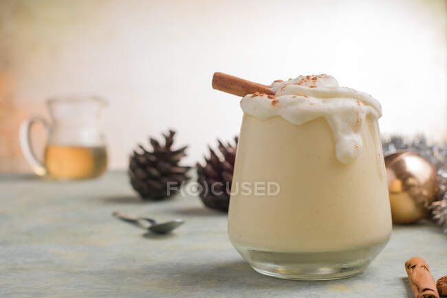 Copo de leite soco com canela em pó no ovo batido branco contra cones de pinho no dia de Natal no fundo claro — Fotografia de Stock