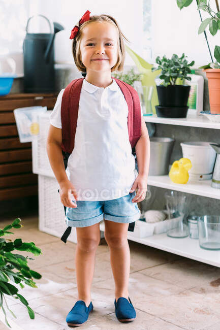 Escolar sonriente con mochila y lazo en el pelo mirando la cámara entre las plantas en maceta en casa - foto de stock