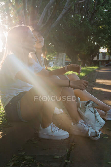 Content best female friends in gumshoes speaking while sitting on pavement border in summer park in sunbeam — Stock Photo