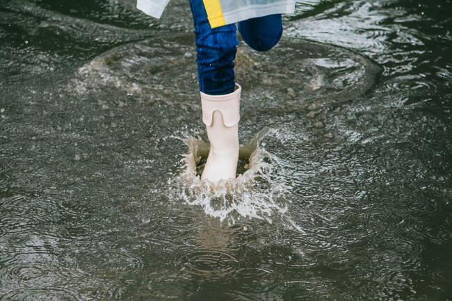 From above crop anonymous child in rubber boots having fun in puddle with splattering aqua on rainy day — Stock Photo