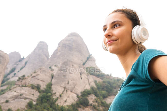 Verträumte junge Touristin schaut weg und genießt den Gesang vom drahtlosen Headset gegen Montserrat und Bäume in Spanien — Stockfoto