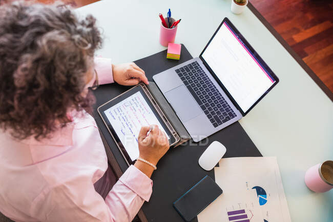 From above of crop anonymous female entrepreneur with tablet and netbook working at desk with graphics on paper sheets — Stock Photo