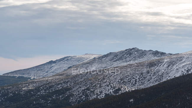 Picturesque scenery of green coniferous forest against snowy mountains under cloudy sky in daytime — Stock Photo