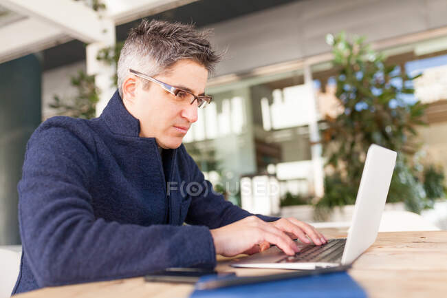 Vista lateral de los jóvenes masculinos concentrados en ropa casual y anteojos escribiendo en el teclado del ordenador portátil mientras está sentado en la mesa en la cafetería al aire libre - foto de stock