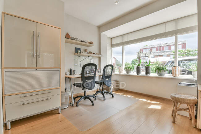 Интерьер современной комнаты с настольным компьютером на столе против кресла и стульев на коврах в светлом доме — стоковое фото