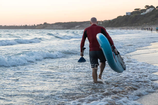 Vista trasera de surfista masculino irreconocible en traje de neopreno llevando tabla de remo mientras camina en la orilla del mar - foto de stock
