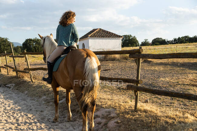 Back view of female riding stallion against stables of riding school in countryside — Stock Photo