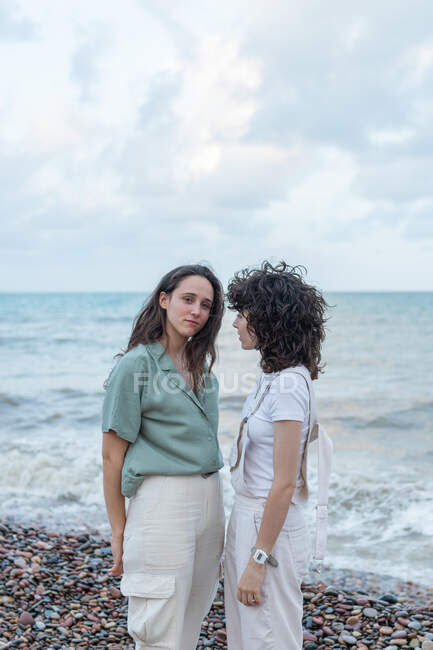 Joven lesbianas amigas en ropa casual de pie mientras mira la cámara en la costa del océano bajo el cielo nublado - foto de stock