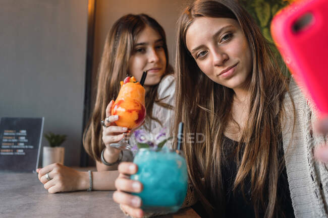 Melhores amigas com copos de bebidas refrescantes tomando auto-retrato no celular à mesa no café urbano — Fotografia de Stock