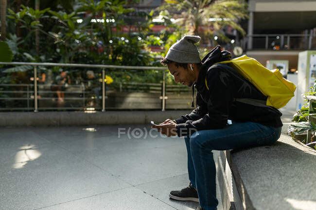 Content African American male hipster with backpack sitting on stone border in street and messaging on mobile phone and listen to music on earbuds — Stock Photo