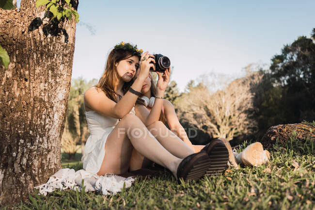 Подросток в сарафане фотографирует парк на профессиональную камеру против неузнаваемой лучшей подруги, сидящей на лужайке — стоковое фото