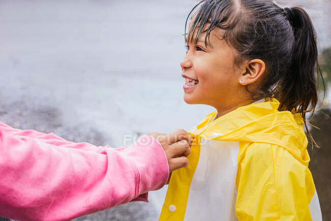 Side view of crop anonymous mom buttoning up slicker on cheerful ethnic girl with wet hair looking away on rainy day — Stock Photo