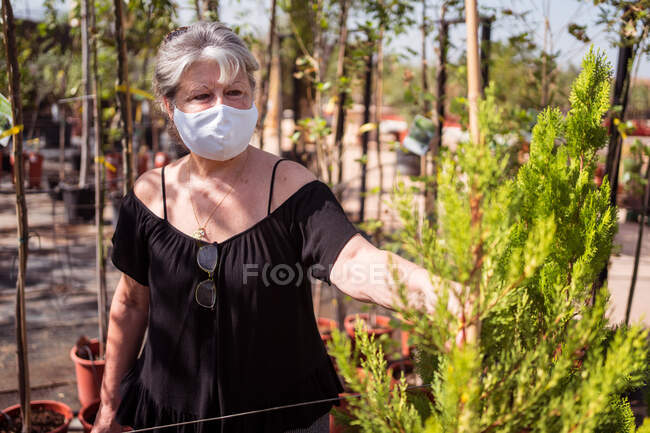 Vista lateral do cliente fêmea maduro na máscara têxtil que escolhe árvores verdes em potenciômetros na loja do jardim no dia ensolarado — Fotografia de Stock