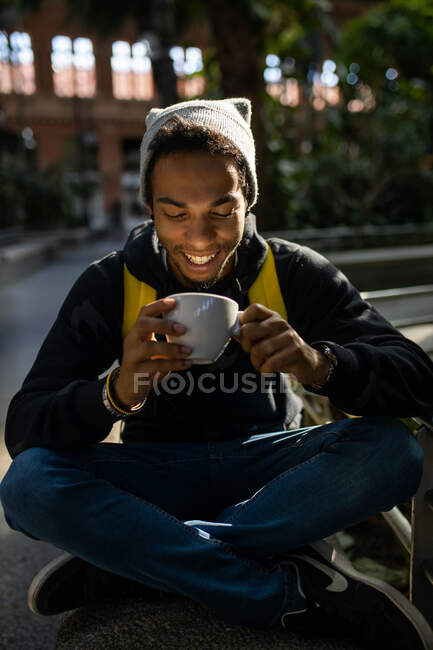 Peaceful smiling African American male hipster with backpack chilling sitting in park and drinking refreshing beverage to go — Stock Photo